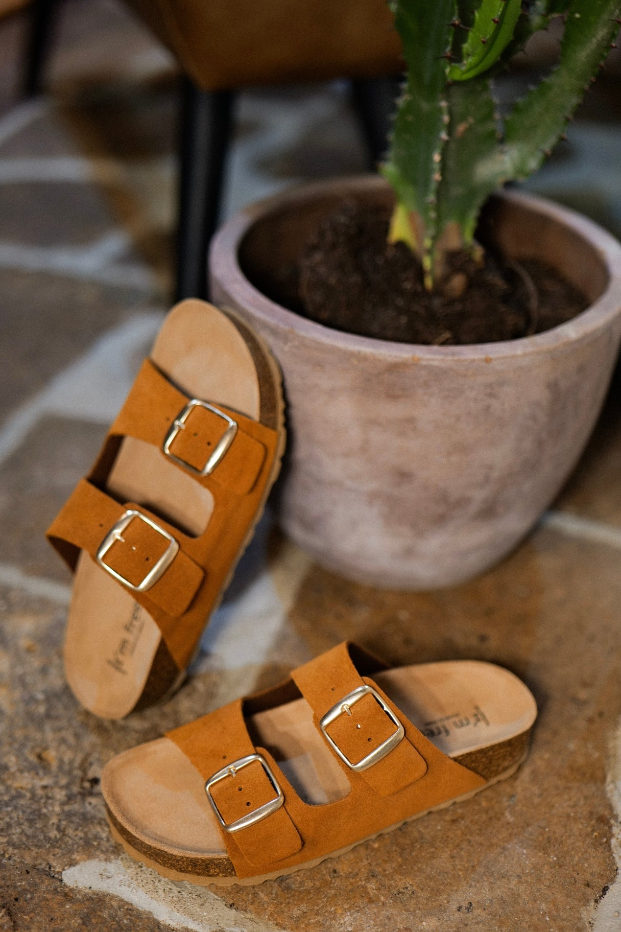 Sandales camel à double boucle en cuir avec semelle effet liège posées devant un pot en terre cuite